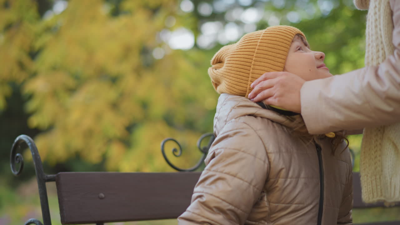 mother lovingly adjusts daughter beanie as child sits calmly on park bench during crisp autumn day with golden trees in background showing warmth care bonding and protection against cold outdoors