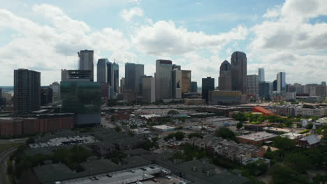 Panoramic-aerial-view-of-downtown-skyscrapers-behind-rush-highway.-Forwards-fly-above-low-buildings.-View-against-bright-clouds-in-sky.-Dallas,-Texas,-US