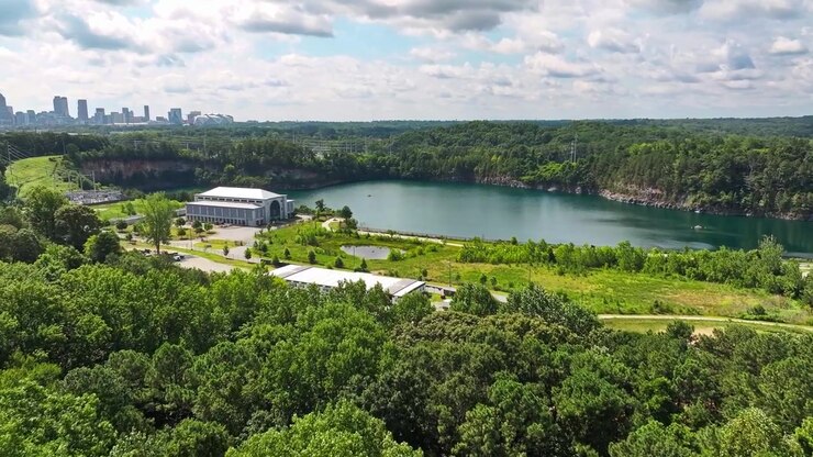 Un lago de cantera turquesa se encuentra junto a un edificio blanco a dos aguas cerca de Atlanta, Georgia; un denso bosque de pinos rodea acantilados rocosos mientras el horizonte del centro de la ciudad se eleva más allá de la línea de árboles. Toma aérea de dron acercándose.