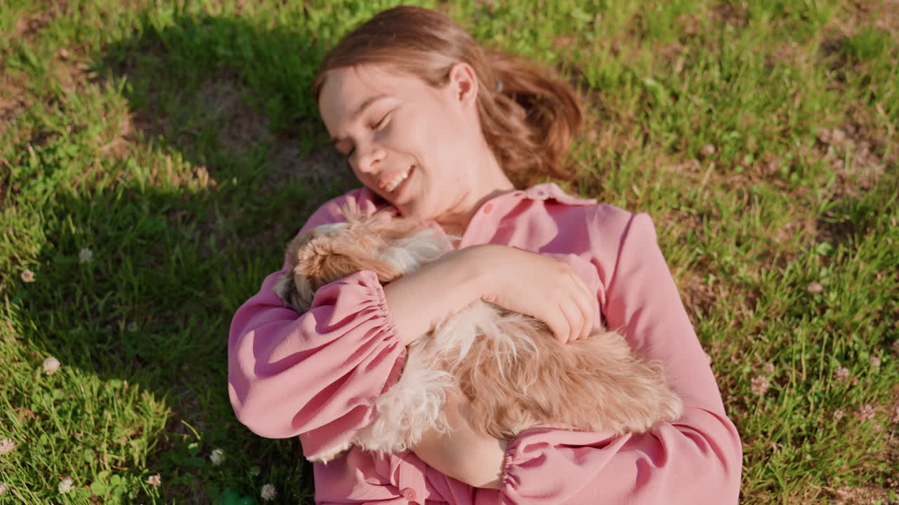 Gentle Woman Embracing Playful Puppy Outdoors, Serene Lady Resting With Fluffy Dog On Bright Grassy Field, Peaceful Woman In Pink Reclines As She Cuddles Her Adorable Puppy Outdoors