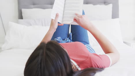 Happy-biracial-woman-lying-on-bed-reading-book-in-sunny-bedroom-at-home,-slow-motion