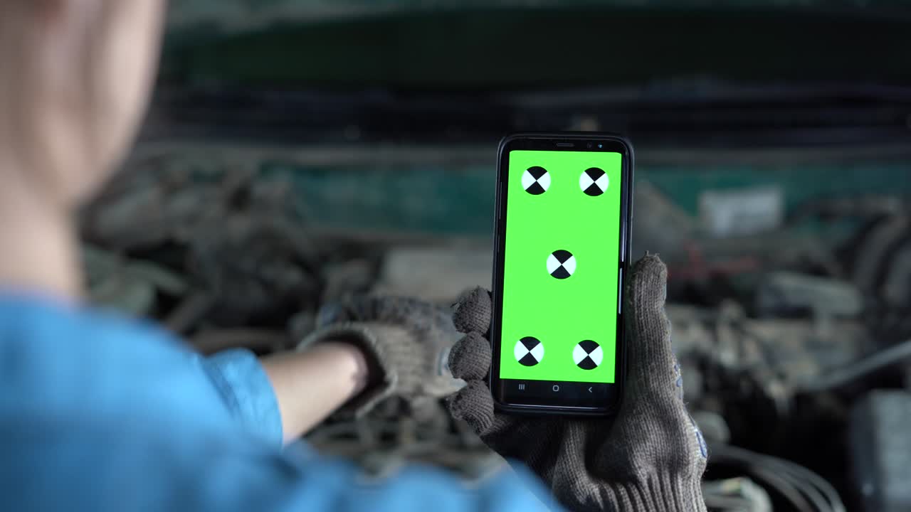 un trabajador del garaje sostiene un teléfono con una pantalla verde frente al capó abierto del coche. reparación de equipos y motor en un taller de automóviles.