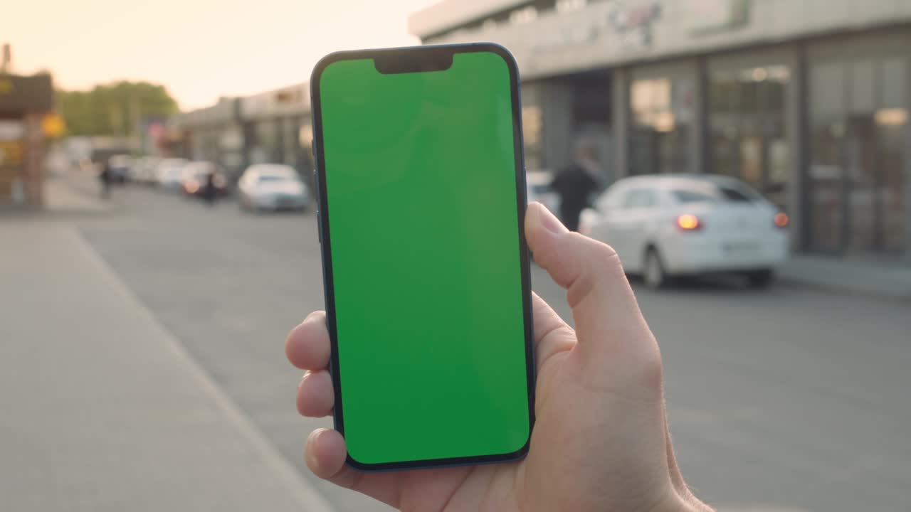 Primo piano della mano di un uomo che tiene uno smartphone con schermo mock-up verde chroma key all'aperto.