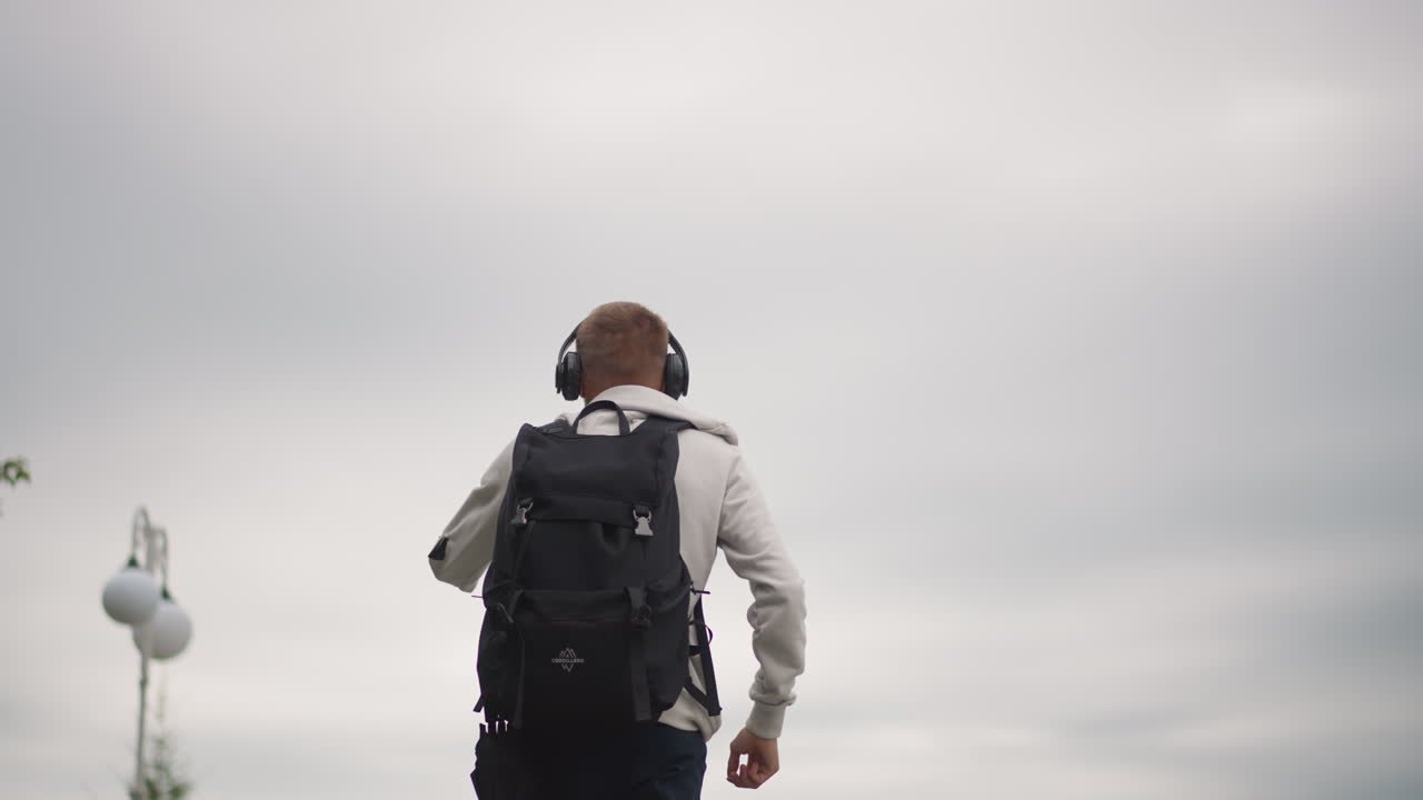 Hombre con mochila bajo un cielo nublado de pie en una pasarela urbana, con las manos en las caderas, escuchando música con auriculares, farola y silueta de árbol, luz nublada y ambiente reflexivo, pausa contemplativa lenta