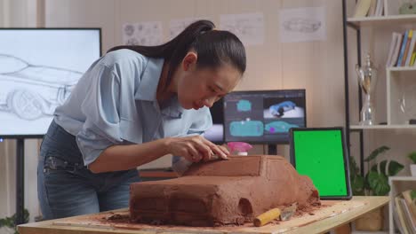 asian woman automotive designer with green screen tablet using rake or wire to smooth out the surface and create details in the sculpture of car clay in the studio