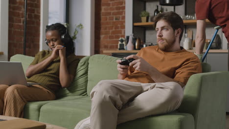 niña afroamericana usando laptop y niño jugando videojuegos sentado en el sofá mientras su compañero de cuarto limpia la casa 1