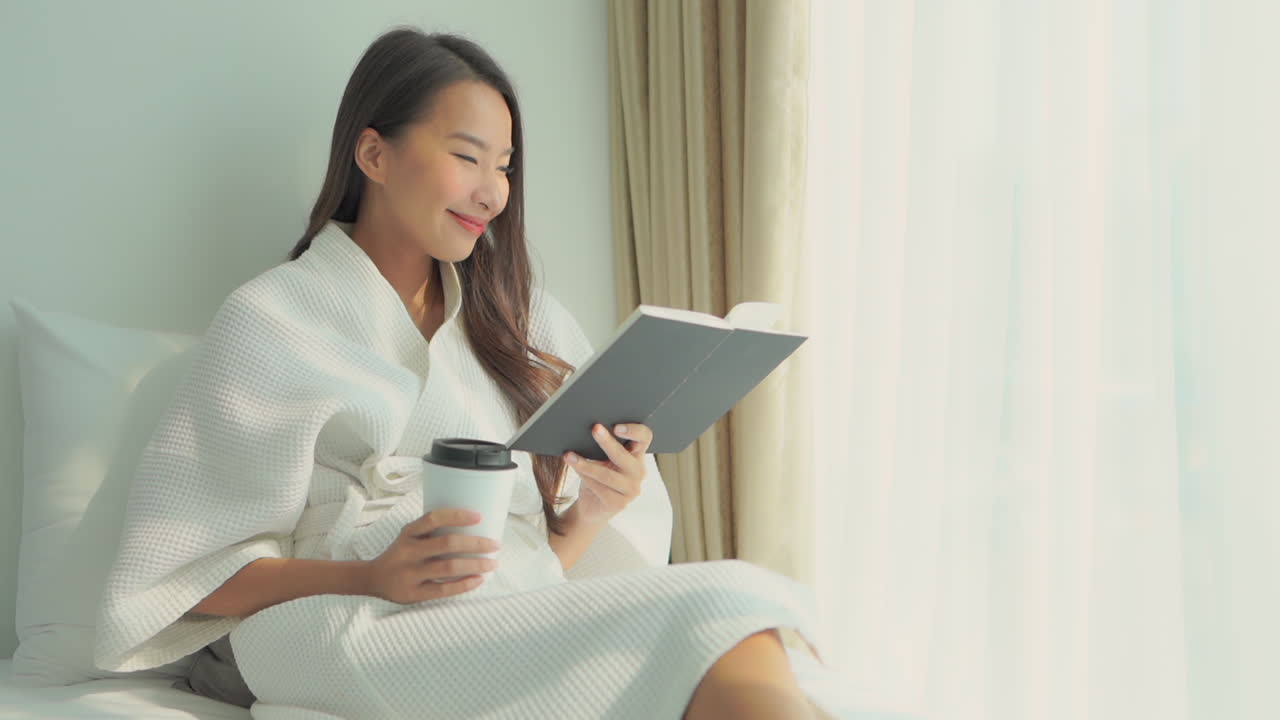 하얀 드레싱 가운을 입고 침대에 누워 있는 동안 창문 근처에서 책을 읽고 커피 한 잔을 들고 있는 젊은 여성