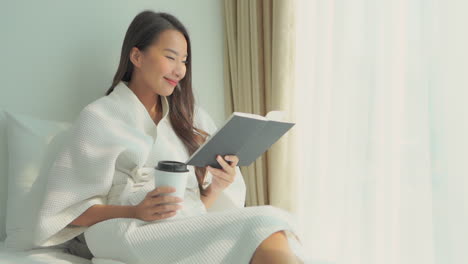 Young-woman-reading-a-book-near-a-window-and-holding-a-cup-of-coffee-while-wearing-a-white-dressing-gown-and-lying-on-a-bed