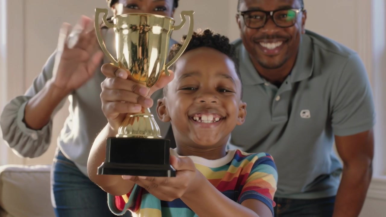 Camera framing boy wearing striped tee holding gold trophy in living room celebrating award
