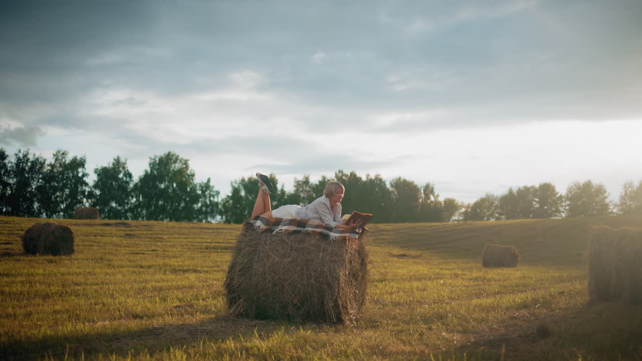 Woman lying on hay bale with checkered blanket, reading book and flipping to a new page under glowing sunlight in serene open field, enjoying peaceful outdoor time