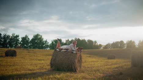 woman lying on hay bale with checkered blanket, reading book and flipping to a new page under glowing sunlight in serene open field, enjoying peaceful outdoor time