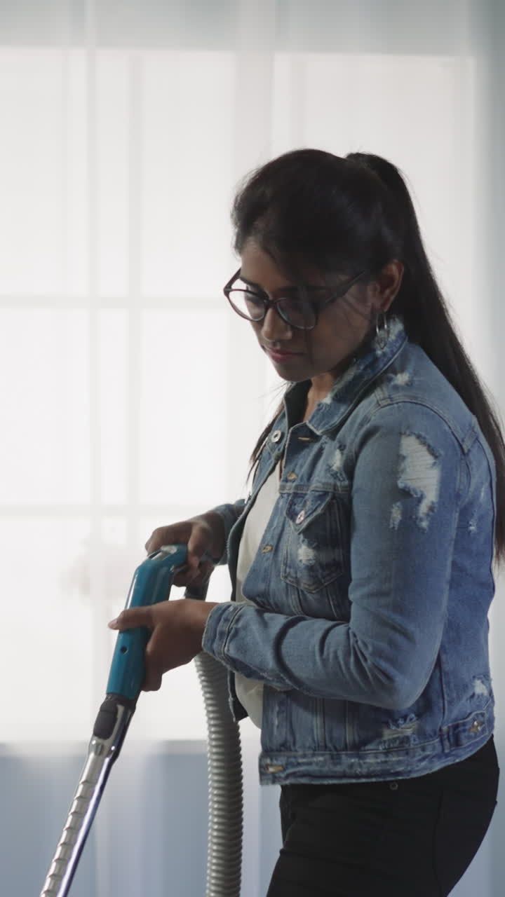 una mujer india enfocada aspira el suelo en la sala de estar contra una ventana brillante y una planta de maceta. una joven ama de casa con gafas limpia la casa la mañana del fin de semana