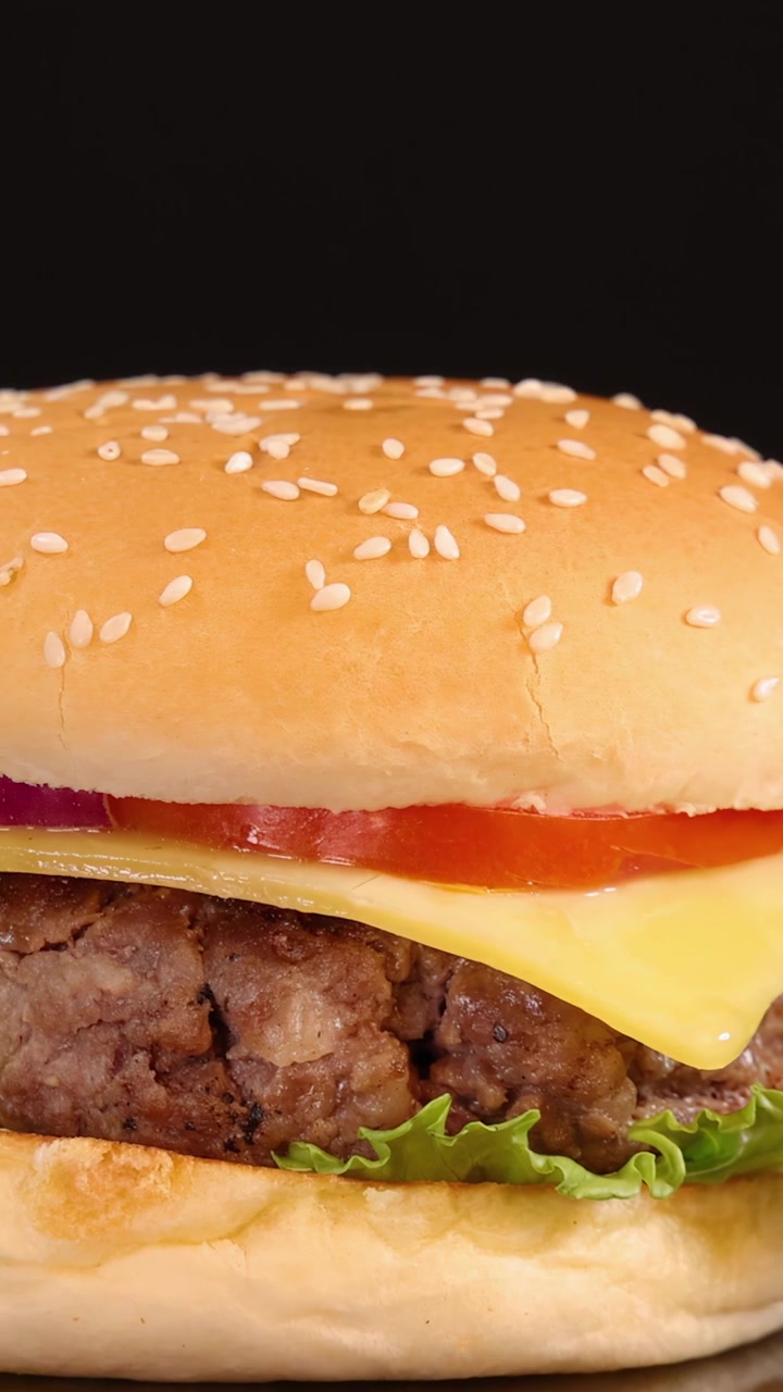 Un classico cheeseburger con lattuga, pomodoro, cipolla e formaggio ruota fluidamente su uno sfondo nero sotto un'illuminazione da studio brillante e uniforme, mettendo in risalto gli ingredienti freschi.