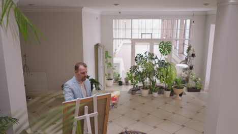 un hombre pensativo dibuja en lienzo en un amplio estudio de arte. un maestro creativo hace obras de arte en un taller decorado con plantas en olla en crecimiento. proceso de terapia artística