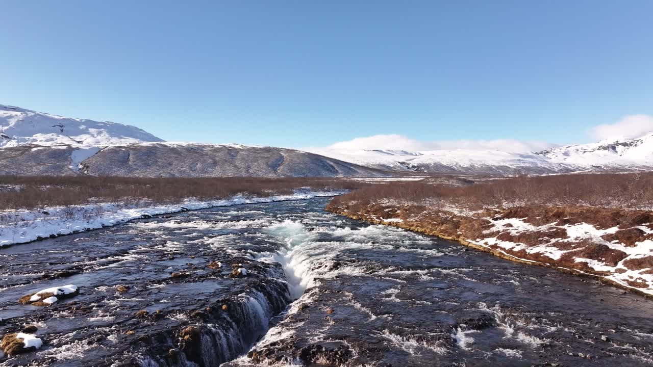 Stunning aerial view of Brúarfoss waterfall near Laugarvatn and Haukadalur, Iceland, icy blue river cascades through rugged winter landscape, surrounded by snow-covered mountains and clear skies.
