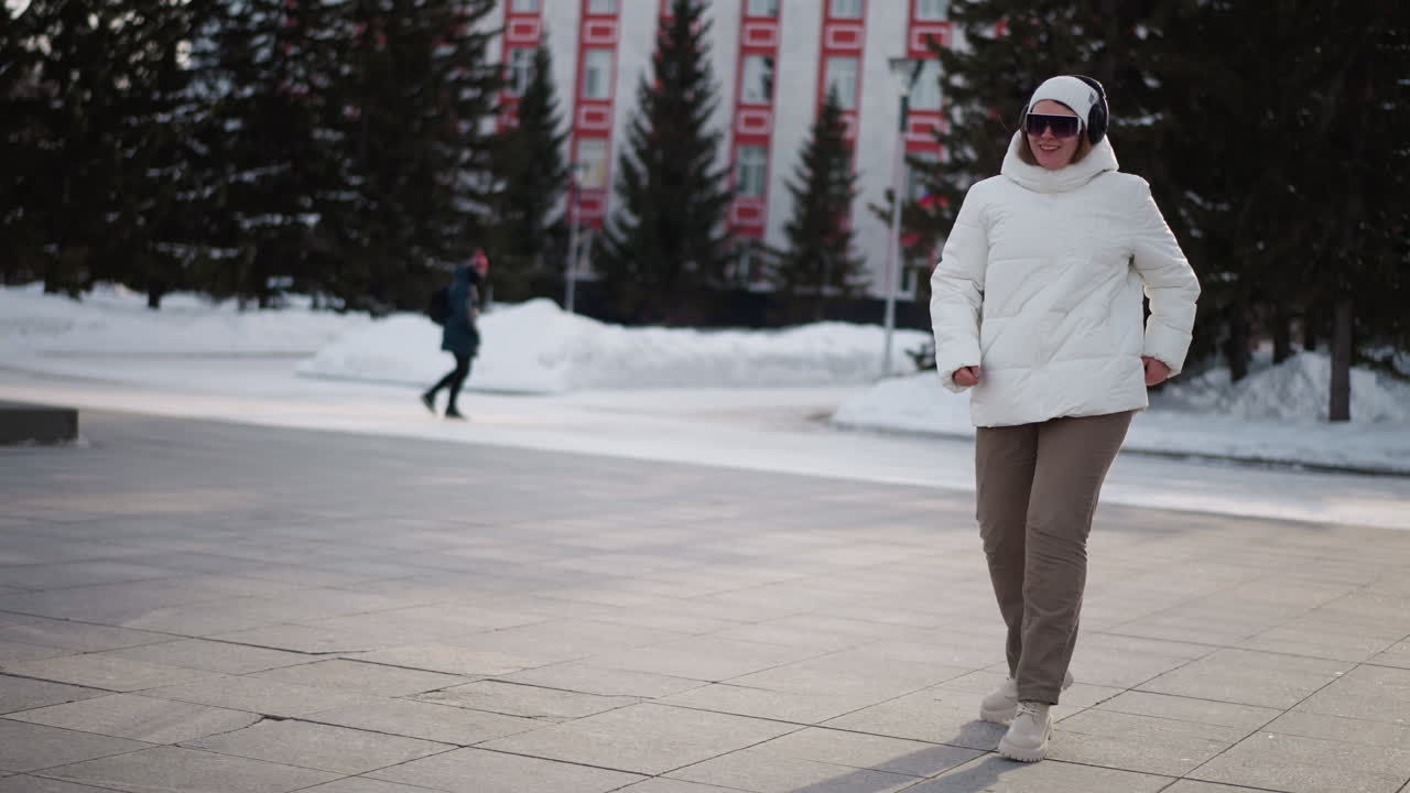 Side view of young dancer wearing white puffer jacket, beanie, goggles and chunky shoes dancing and jumping on snow dusted plaza while people walk in background under pale winter light