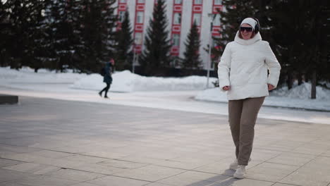 side view of young dancer wearing white puffer jacket, beanie, goggles and chunky shoes dancing and jumping on snow dusted plaza while people walk in background under pale winter light