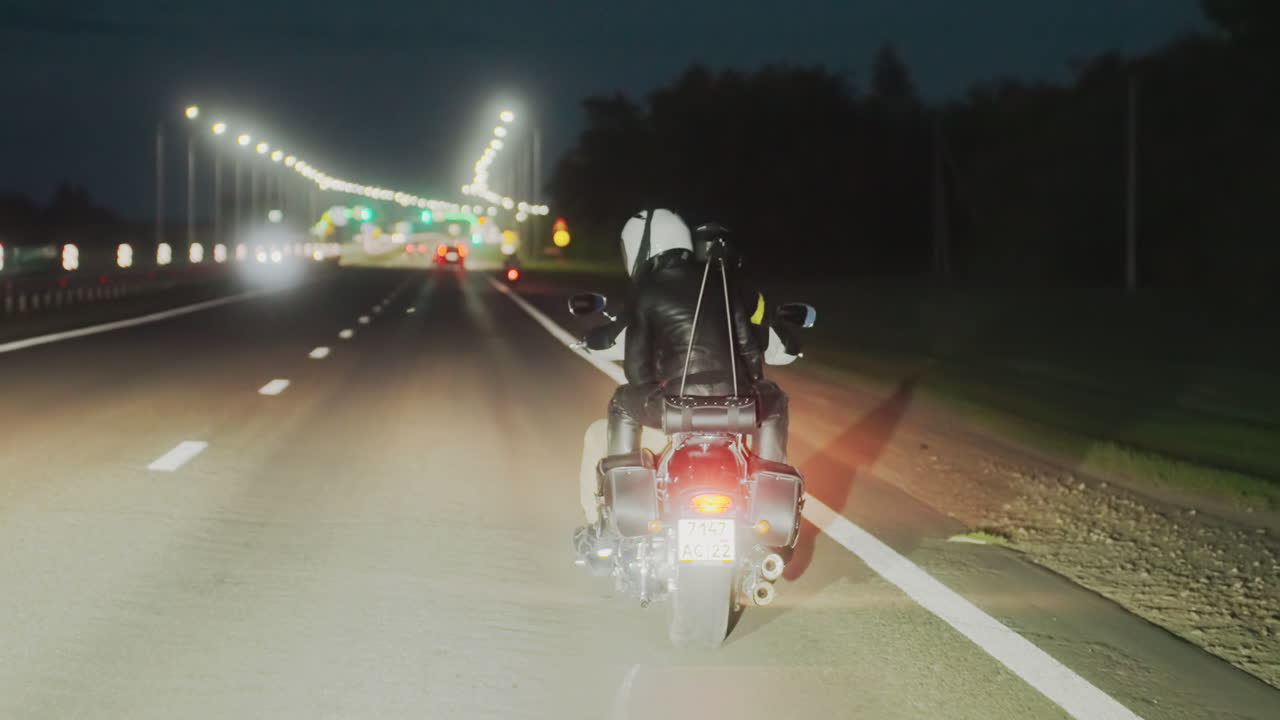 Back view of motorcycle rider wearing helmet and leather jacket traveling on illuminated highway at night, tail light glowing, moving toward city with visible traffic lights and cars ahead