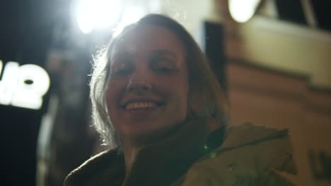 Smiling-pretty-girl-looks-up-at-the-luminous-sign-board-then-down-to-the-camera.-Night-time.-Light-behind.-Shooting-from-below