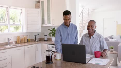 happy african american gay male couple sitting at countertop in kitchen, using laptop, slow motion
