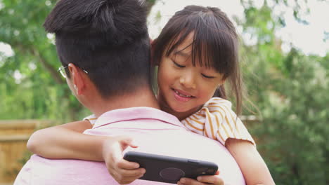 Asian-Father-Cuddling-Girl-In-Garden-As-Girl-Looks-Over-His-Shoulder-At-Mobile-Phone