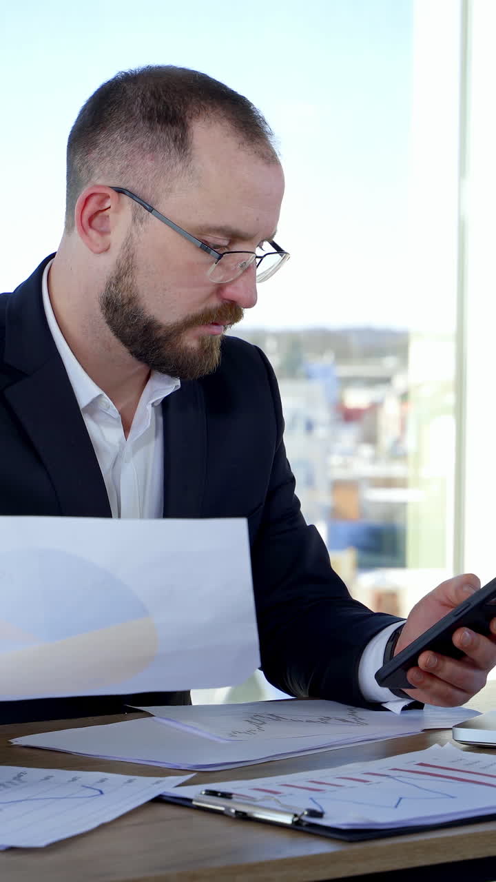 Serious businessman in suit working in office. Entrepreneur sitting at table using laptop, phone and project papers on window background. Vertical video