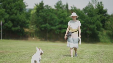 a middle-aged casually looking woman, wearing a handmade knitted dress and a wide-brimmed sun hat, enjoying the walk in the town park with her dog