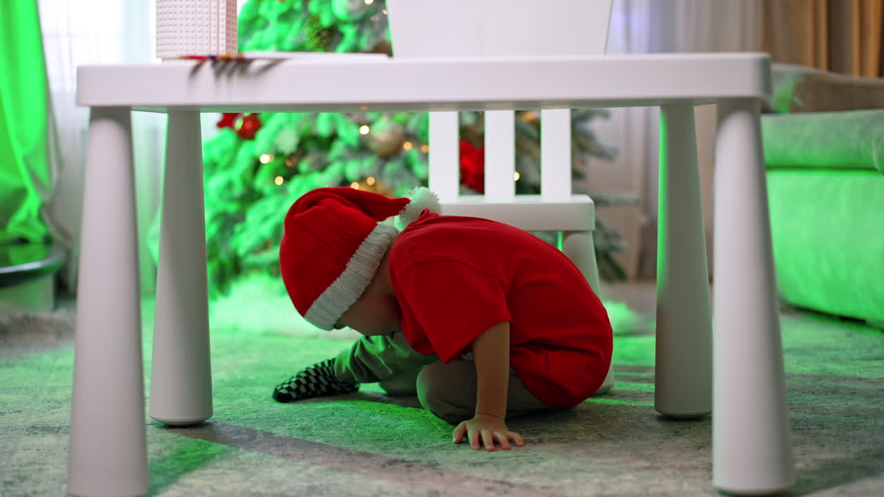 Caucasian toddler in red t-shirt and Santa cap crawls under the desk. Little kid having fun at home for Christmas.