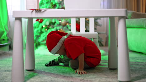 caucasian toddler in red t-shirt and santa cap crawls under the desk. little kid having fun at home for christmas.