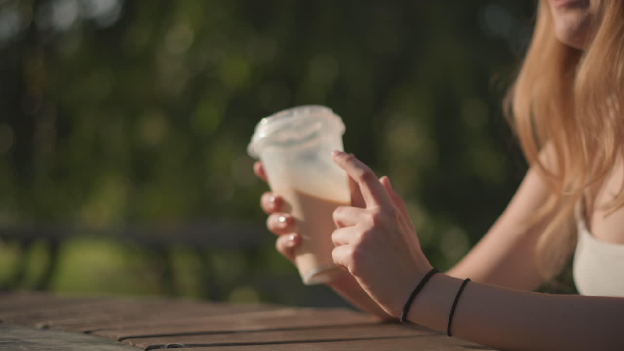 Giovane donna che tiene in mano un latte macchiato freddo all'aperto su un tavolo di legno, luce del sole che filtra tra gli alberi, primo piano delle mani che sorreggono la tazza con la schiuma, abbigliamento estivo casual, atmosfera rilassata e spontanea, atmosfera da bar nel parco