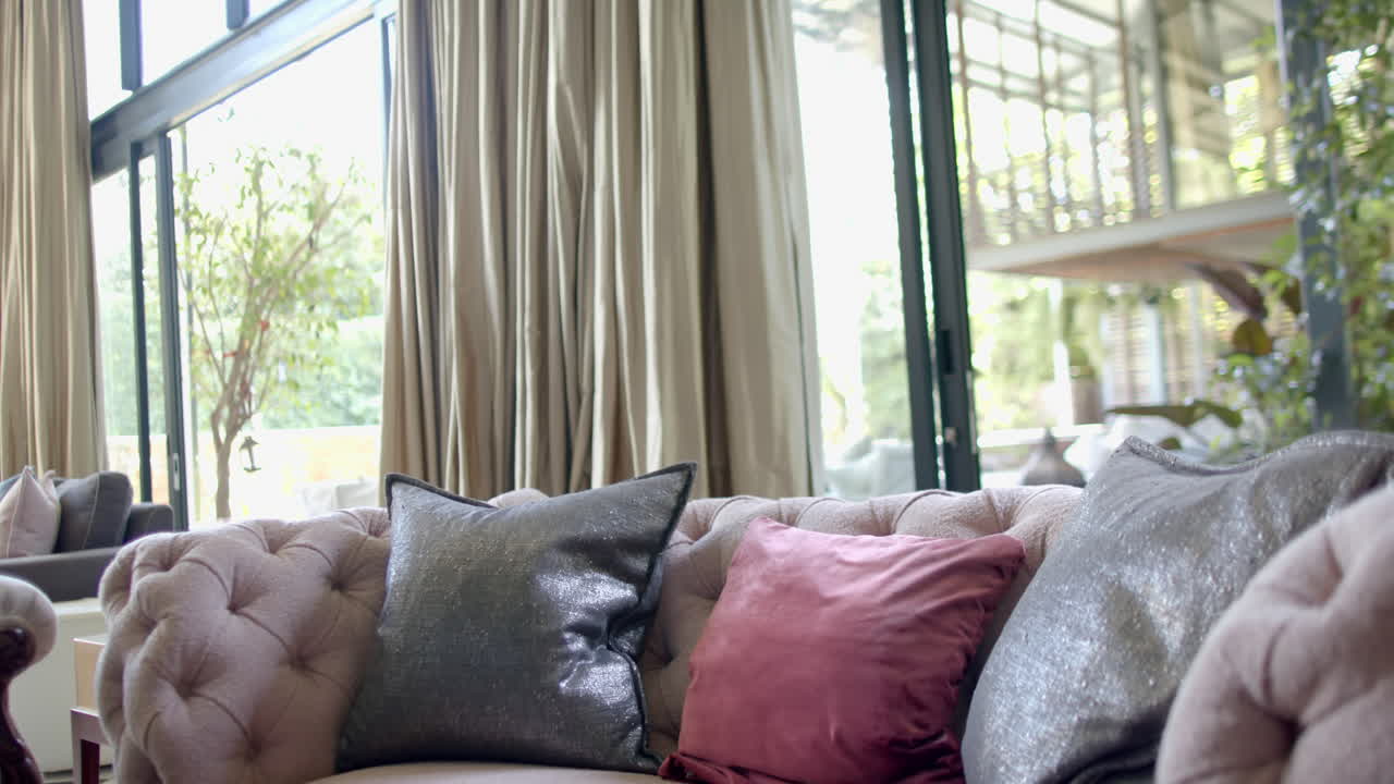 sofá beige con almohadas, cortinas de oro y ventanas enormes en la sala de estar soleada, cámara lenta