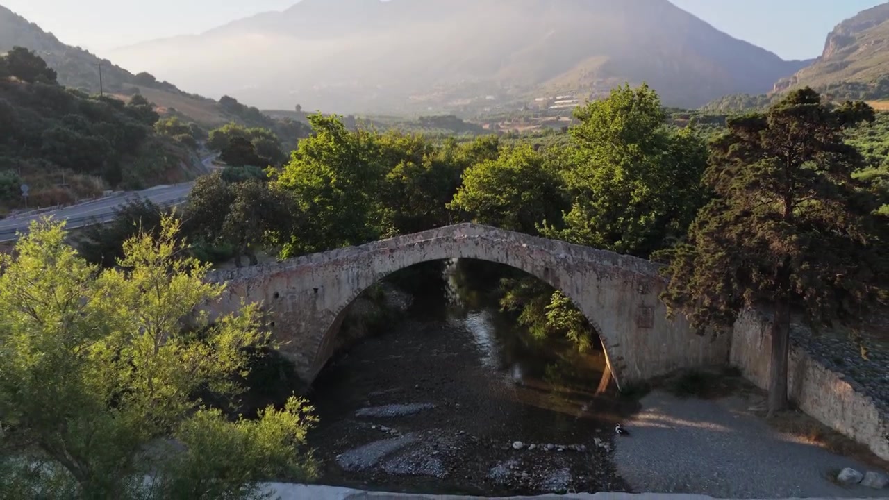 Vista aerea con drone del vecchio arco in pietra del Ponte di Preveli che attraversa un letto di fiume roccioso fiancheggiato da alberi, Preveli, Grecia