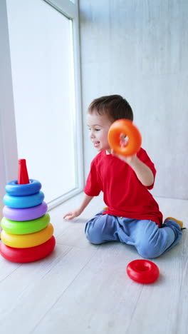 cheerful baby boy sitting on the floor near the window builds a pyramid. joyful toddler having fun at home. vertical video.