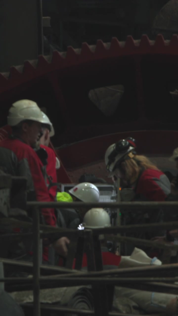 Workers In Helmets Repair Giant Gear, Tense Focused Maintenance Inside Dim Industrial Mill With Scaffolding,