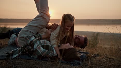 A-little-blonde-girl-in-a-white-jacket-runs-to-her-parents,-a-brunette-man-and-a-girl-in-a-green-checkered-shirt-jumps-on-them-and-knocks-them-onto-the-mat-during-a-picnic-outside-the-city-on-a-summer-evening