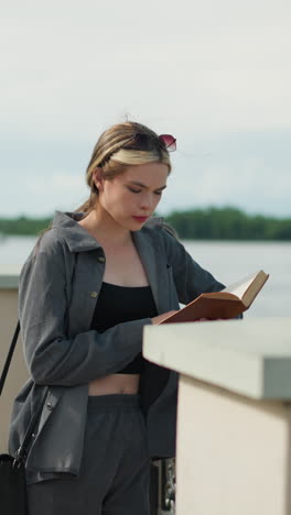 mujer con ropa gris apoya su mano derecha en una valla de hierro mientras lee un libro a la orilla del río, el viento sopla suavemente, con árboles y el agua visibles en el fondo