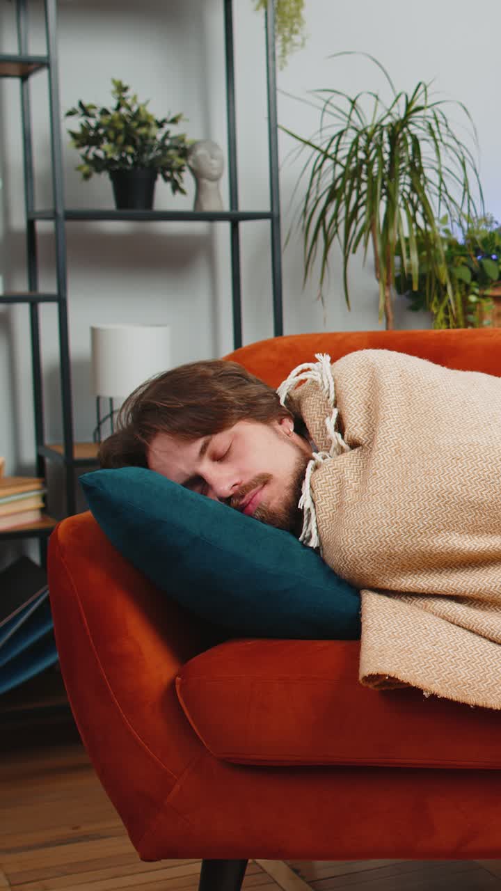 Tired young man lying down on comfortable couch taking a rest daytime nap relaxing falling asleep