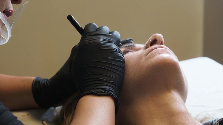 Eyebrow mapping shows a beauty technician holding a precision marking tool while stabilizing client's brow area with black gloves during cosmetic tattoo preparation. Eyebrow mapping, Brow shaping,