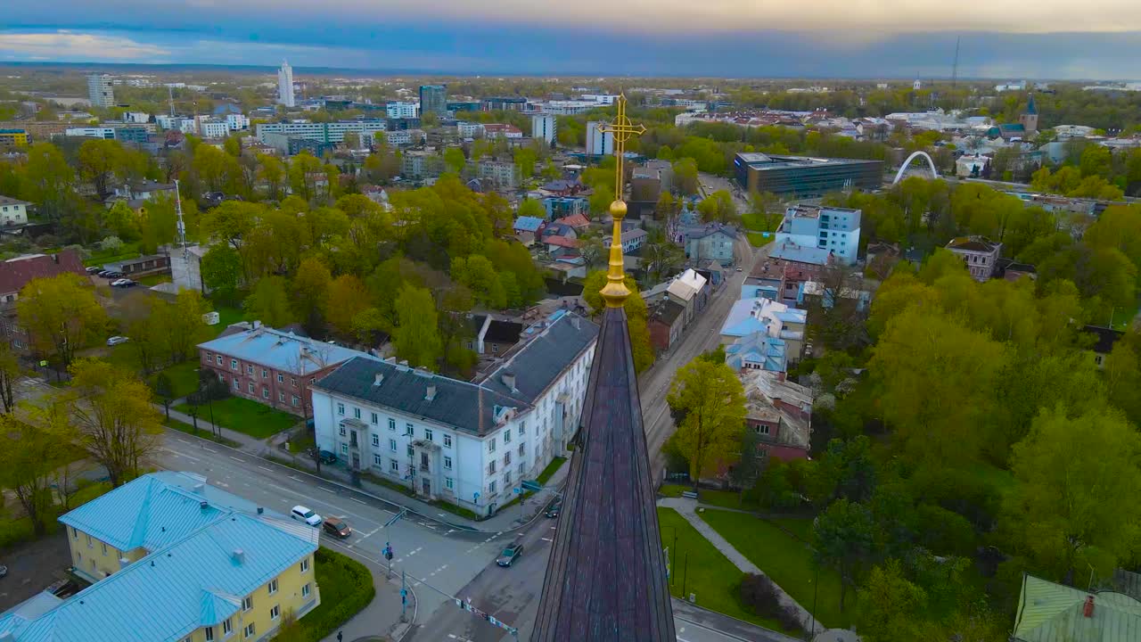 Aerial drone footage orbiting and spinning around a golden colored shiny holy cross on top of a church tower in Tartu Estonia during evening or morning time. Green trees and grass in urban area.