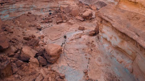 two adventure seekers make their way across a rocky canyon filled with debris and rocks. the desert terrain features patches of clay soil, red rocks, and a hint of dried-out watering holes.