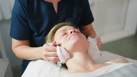 under bright lamp light skin care expert cleans white woman face with damp cotton pad during facial prep in modern clinic room illuminated by clinical lamp creating calm hygienic atmosphere