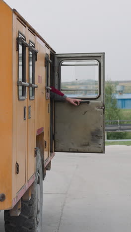 workers in uniform get in big orange vehicle closing doors preparing to drive to mine. heavy mining machinery parked on street on cloudy day