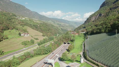 highway going through the mountain next to the river, cars going to the tunnel, going up drone view, sunny weather