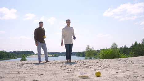 zwei kaukasische junge mannfreunde, die an einem sonnigen tag am strand boccia spielen