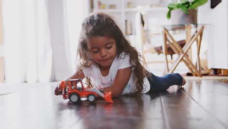 young hispanic girl lying on the floor in the sitting room playing with toy digger truck, side view