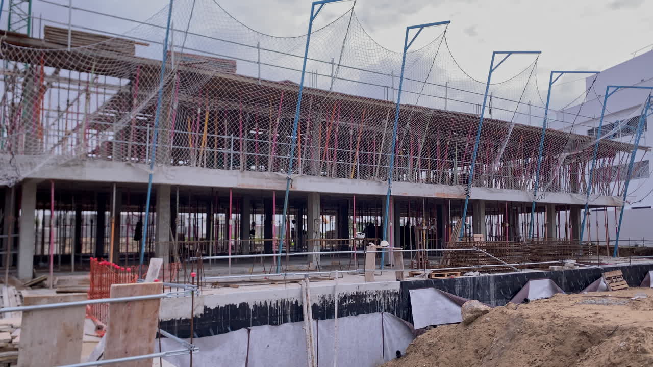 Proyek konstruksi bangunan dengan struktur beton, perancah, dan jaring pengaman di bawah langit berawan. Mewakili arsitektur dan pembangunan