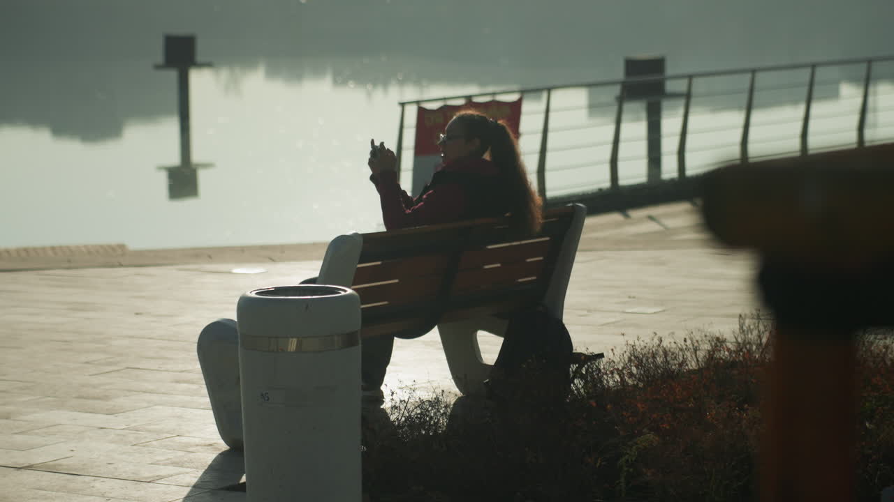 Person on bench by waterfront, hooded silhouette facing misty pier early morning mood with lifebuoy and railing, muted tones, contemplative posture suggesting commuter pausing between journeys