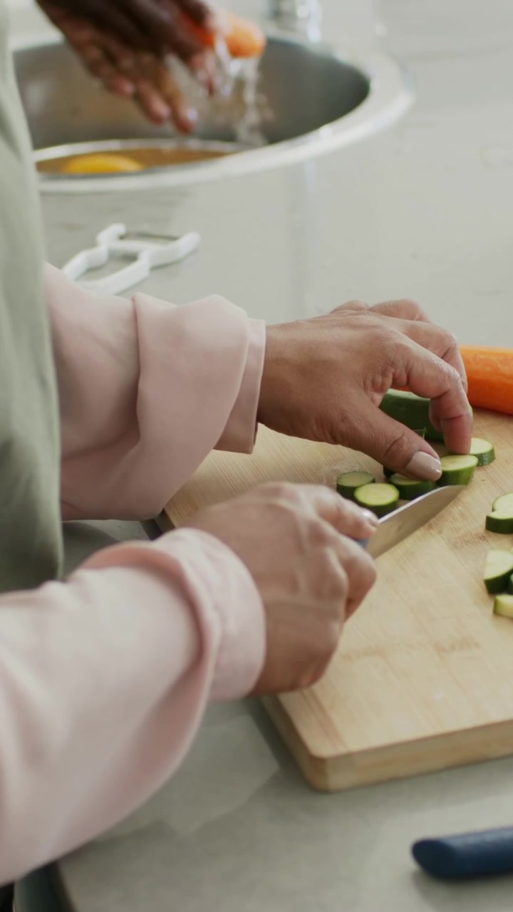 Video verticale: Iniziando la preparazione, donna con grembiule verde che affetta zucchine su un tagliere, uomo che sciacqua una carota.