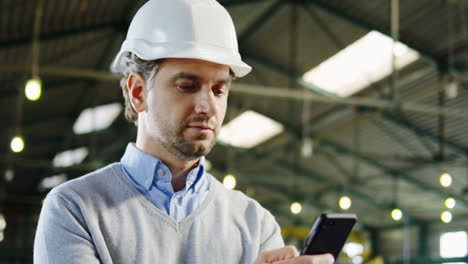 close-up view of caucasian male worker wearing a helmet typing on smartphone in a factory