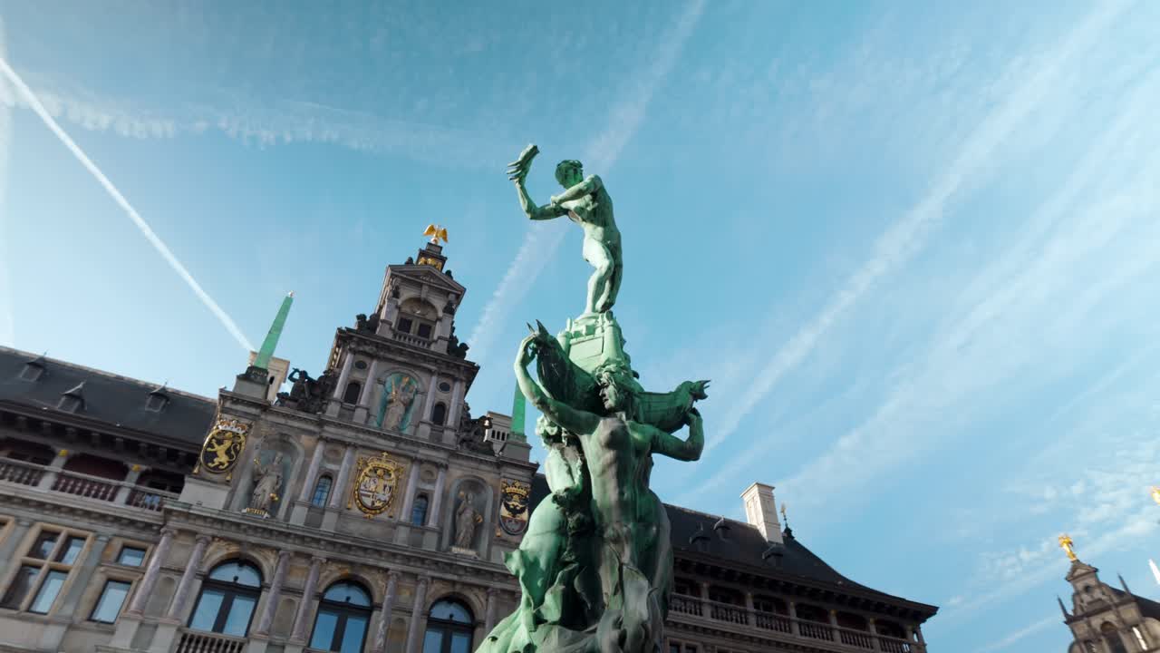 Low-angle view of the iconic Brabo Fountain statue in the Grote Markt of Antwerp, Belgium. Historic guildhalls and the City Hall frame the legendary bronze sculpture under a blue sky